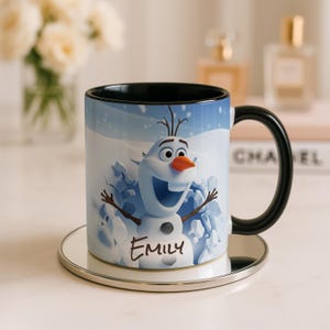 May include: A ceramic mug featuring a cartoon snowman character with a carrot nose and stick arms, set against a snowy background. The mug has a black handle and rim, and the name "Emily" is written on the front.