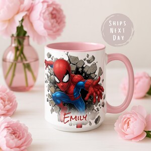 K&ouml;nnte beinhalten: Wei&szlig;e Keramik-Tasse mit Spider-Man, der durch eine Wand bricht. Die Tasse hat einen rosa Innenraum und Henkel, mit dem Namen "Emily" in Rot. Umgeben von rosa Blumen.
