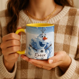 May include: White ceramic mug with a yellow handle and interior. The mug features a cartoon image of Olaf the snowman from Frozen, with the name "Emily" written below. The mug is held by a person.