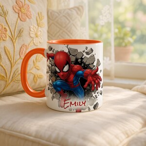 K&ouml;nnte beinhalten: Wei&szlig;er Keramikbecher mit orangefarbenem Innenraum und Henkel, mit einem Spiderman-Motiv. Das Design zeigt Spiderman, der durch eine Wand bricht, mit dem Namen "EMILY" darunter. Der Becher steht vor einem weichen, neutralen Hintergrund.