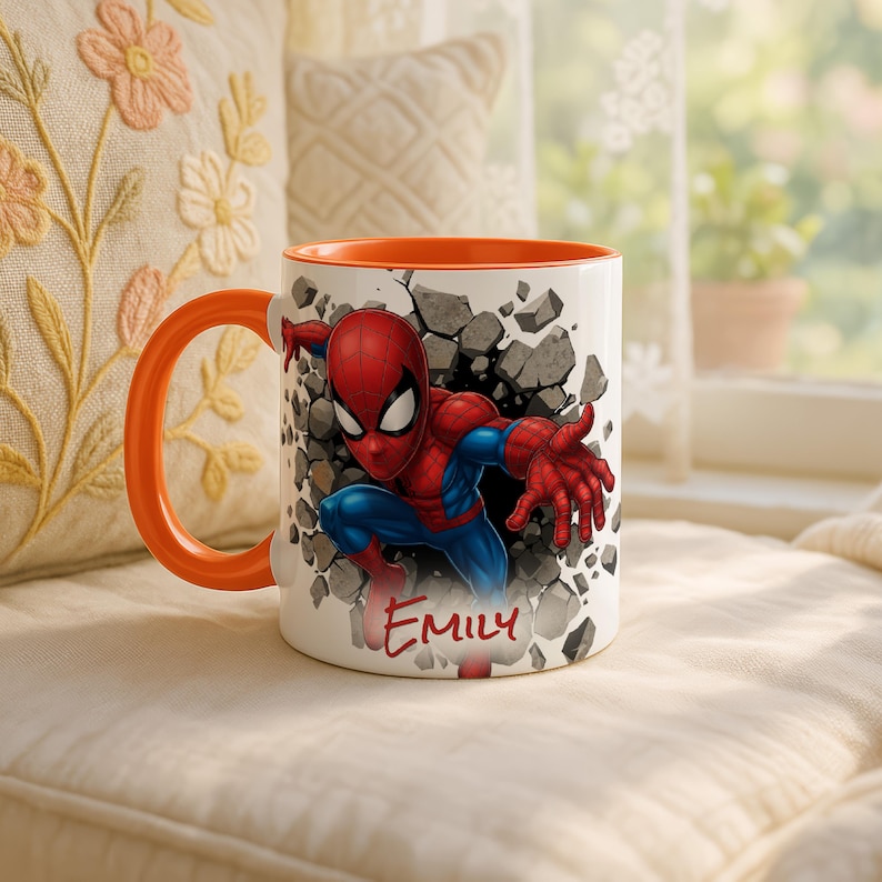 K&ouml;nnte beinhalten: Wei&szlig;er Keramikbecher mit orangefarbenem Innenraum und Henkel, mit einem Spiderman-Motiv. Das Design zeigt Spiderman, der durch eine Wand bricht, mit dem Namen "EMILY" darunter. Der Becher steht vor einem weichen, neutralen Hintergrund.