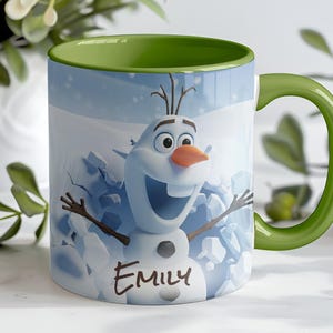 May include: A ceramic mug featuring Olaf from Frozen, with a green handle and interior. The mug has a light blue background with a snow scene and the name "Emily" written on it. The mug is decorated with a smiling Olaf.