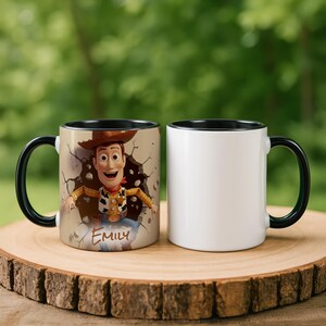 May include: Two ceramic mugs with black handles and rims. One mug features a colourful image of the character Woody from Toy Story, breaking through a wall, with the name "Emily" below. The other mug is plain white.