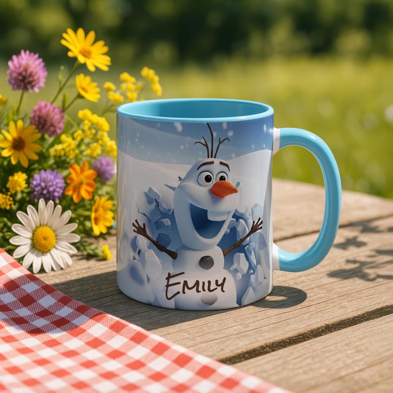 May include: A ceramic mug featuring Olaf the snowman from Frozen, with a light blue handle and interior. The mug has the name "Emily" printed on it. The background shows a wooden table, flowers, and a red and white checkered cloth.