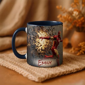 May include: A ceramic mug with a dark blue handle and interior. The exterior features a graphic of Iron Man breaking through a wall, with the name "Emily" in red script. The mug is set on a beige knitted surface.