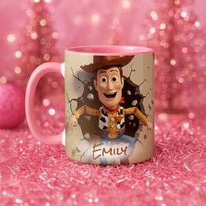 May include: A pink and beige ceramic mug featuring a cartoon character breaking through a wall. The character is wearing a cowboy hat, vest, and blue jeans. The name "Emily" is written on the mug.