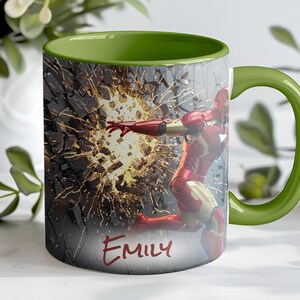 May include: A ceramic mug with a green interior and handle, featuring an illustration of Iron Man breaking through a wall. The design includes the name "Emily" in red script. The mug is white with a wraparound design.