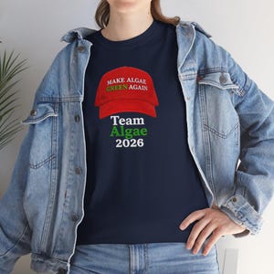 May include: A navy blue t-shirt featuring a graphic of a red baseball cap with the text "MAKE ALGAE GREEN AGAIN" in white and green, followed by the text "Team Algae 2026" in white and green, worn under a light blue denim jacket.