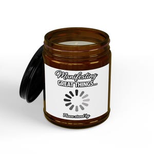 May include: A brown glass jar candle with a black lid. The candle has a white label with the text "Manifesting Great Things..." and a loading symbol. The bottom of the label reads "Please stand by."
