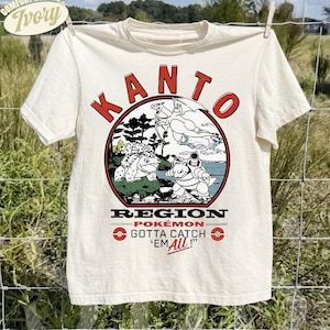 Può includere: Una t-shirt a maniche corte color avorio con una stampa grafica che riporta il testo KANTO REGION POKEMON GOTTA CATCH 'EM ALL! Il design include illustrazioni di Charizard, Venusaur e Blastoise all'interno di una cornice circolare con accenti rossi.