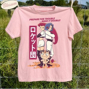 Könnte beinhalten: Ein hellrosa T-Shirt mit kurzen Ärmeln und einer Grafik von Jessie, James und Mauzi aus Team Rocket. Der Text lautet PREPARE FOR TROUBLE...MAKE IT DOUBLE! und enthält japanische Schriftzeichen in einem vertikalen rosa Rechteck links.