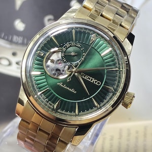 May include: A gold-tone Seiko automatic watch with a metallic green sunray dial. The watch features a gold-tone link bracelet, a circular case, gold-tone hour markers, and a visible open-heart balance wheel. The dial includes a 24-hour sub-dial.