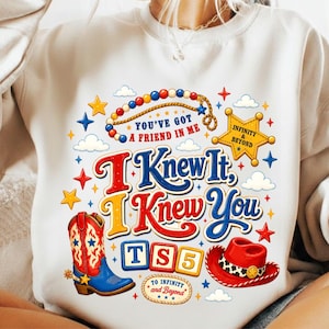 K&ouml;nnte beinhalten: Ein cremefarbenes Sweatshirt mit Rundhalsausschnitt und einem bunten Grafikdruck, der einen Cowboystiefel, einen roten Hut, einen Sheriffstern und Alphabet-Bl&ouml;cke mit der Aufschrift TS5 zeigt. Der Text lautet You've Got A Friend In Me, I Knew It, I Knew You und To Infinity And Beyond.