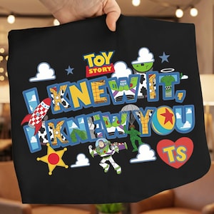 K&ouml;nnte beinhalten: Ein schwarzes T-Shirt mit einer bunten Toy Story-Grafik und dem Text "I KNEW IT, I KNEW YOU" sowie "TS" in einem roten Herz. Das Design enth&auml;lt Illustrationen von Buzz Lightyear, einem gr&uuml;nen Spielzeugsoldaten mit Fallschirm, einer Rakete und einem Ball.