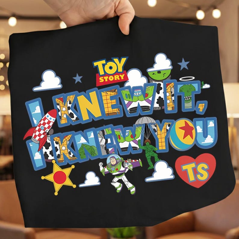 K&ouml;nnte beinhalten: Ein schwarzes T-Shirt mit einer bunten Toy Story-Grafik und dem Text "I KNEW IT, I KNEW YOU" sowie "TS" in einem roten Herz. Das Design enth&auml;lt Illustrationen von Buzz Lightyear, einem gr&uuml;nen Spielzeugsoldaten mit Fallschirm, einer Rakete und einem Ball.