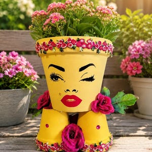 Puede incluir: Una maceta de terracota amarilla con un rostro pintado, pestañas negras, cejas y labios rojos. El borde está decorado con pequeñas flores rosas. Dos macetas amarillas más pequeñas sirven de base, adornadas con flores rosas y pedrería.
