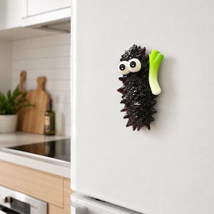 Könnte beinhalten: Ein dunkelbrauner, texturierter Kühlschrankmagnet in Form einer Seegurke mit zwei großen weißen Wackelaugen und einer kleinen grünen Frühlingszwiebel an der Seite. Das Objekt ist ein unebener, länglicher Zylinder mit einer dunklen, glänzenden Oberfläche.