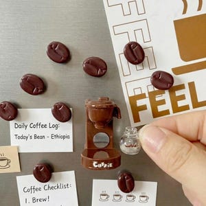 Könnte beinhalten: Ein Set aus braunen Kaffeebohnen-Magneten, ein Miniatur-Kaffeemaschinen-Magnet mit der Aufschrift "Coffie" und ein kleiner Magnet in Form einer klaren Glaskaraffe. Weiße Papiernotizen mit dem Text "Daily Coffee Log" haften auf der Fläche.