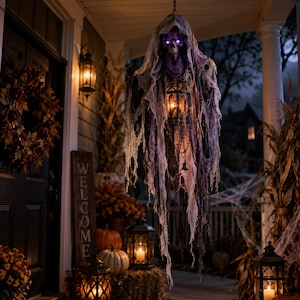 Könnte beinhalten: Eine hängende Halloween-Dekoration mit einer Skelettfigur, die in zerfetzten violetten und grauen Stoff gehüllt ist. Die Figur hat leuchtend violette Augen und hält eine dunkle Metalllaterne mit warmem Licht, die an einer Kette hängt.