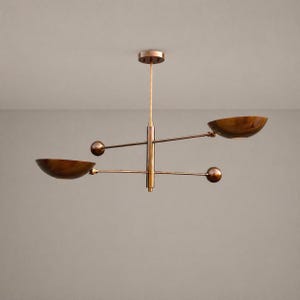 Peut inclure: Un lustre moderne de couleur cuivrée, avec une tige centrale verticale et des bras horizontaux. Deux abat-jours en forme de bol et deux sphères ornent les bras. Le luminaire est suspendu au plafond par une fine tige.