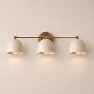 May include: A three-light wall fixture featuring a horizontal brass bar and a circular brass wall plate. Three matte cream-colored dome-shaped shades are attached to the bar, each housing a single light bulb that emits a warm glow downward.