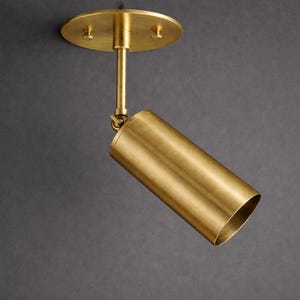 May include: A brass-colored, cylindrical spotlight fixture. The light is mounted on a round ceiling plate with a straight rod and a pivoting joint. The spotlight is angled downward, showcasing its open end. The fixture is designed for directional lighting.