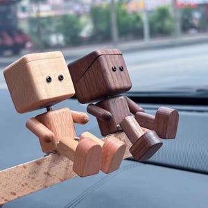 May include: Two wooden car dashboard ornaments shaped like small figures. One is light-colored, the other is dark brown. Each has a box-shaped head with two black dot eyes, arms, legs, and feet. They are sitting on a wooden plank.