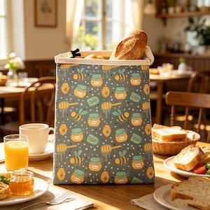 Puede incluir: Una bolsa de pan gris con un patrón repetido de tarros de miel, abejas y cucharones de miel. La bolsa está llena de pan. La bolsa está sobre una mesa de madera con jugo de naranja y un plato de comida.
