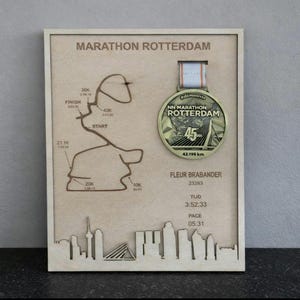 Op de afbeelding: Houten ingelijste display met een Marathon Rotterdam medaille. De lijst bevat een lasergesneden kaart van de race, een silhouet van de skyline en details van de loper. De medaille is goudkleurig met een wit en oranje lint.