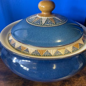 May include: A blue ceramic casserole dish with a lid. The lid has a decorative band with geometric patterns in brown, white, and green. The knob on the lid is light brown. The dish has a glossy finish. The background is blue.