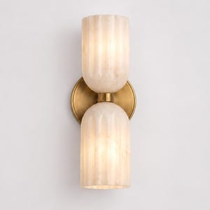 May include: A wall sconce featuring two vertically stacked, fluted, translucent white alabaster glass shades. The shades are connected by a central brushed brass cylindrical component, which is mounted to a circular brushed brass wall plate.