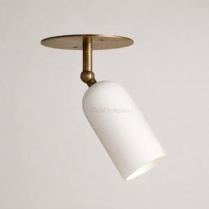 May include: A ceiling-mounted spotlight featuring a circular brass base and a cylindrical white glass shade. The fixture includes an adjustable brass swivel joint connecting the base to the shade, with the text CalaDesignss printed on the glass.