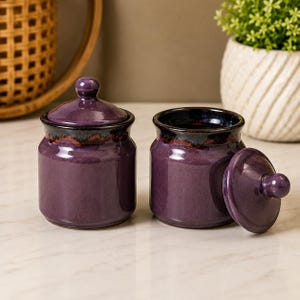May include: Two cylindrical ceramic jars with a glossy purple glaze. Each jar features a dark, mottled rim with hints of black and brown. One jar has a matching purple lid placed on top, while the other jar has its lid resting beside it.