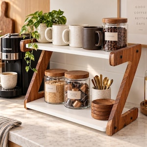 May include: A two-tier countertop organizer with white shelves and Z-shaped wooden side supports. The top shelf holds two speckled mugs, a dark brown mug, and a glass jar labeled COFFEE. The bottom shelf holds glass jars labeled SUGAR and COFFEE PODS, a speckled utensil holder with gold spoons, and a stack of small wooden coasters.