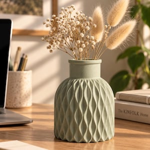May include: A sage green ceramic vase with a textured, vertical wave pattern on its rounded body. The vase contains a dried floral arrangement featuring small white baby's breath flowers and three fluffy, light beige bunny tail grasses.