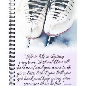 Op de afbeelding: Een spiraalgebonden notitieboek met een paar witte schaatsen met zilveren messen. Het notitieboek bevat een citaat over het leven en schaatsen. De schaatsen staan op een besneeuwd oppervlak.