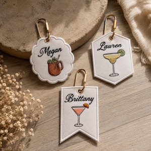May include: Three white embroidered fabric tags with gold-tone metal lobster clasps. One scalloped tag features the name Megan and a brown mug. One hexagonal tag features the name Lauren and a yellow cocktail. One pennant tag features the name Brittany and a pink cocktail.