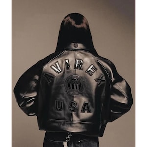 May include: Black leather jacket with the word "AVIREX" and "USA" stitched on the back. The jacket has a round emblem in the center. The jacket is worn by a person with dark hair.