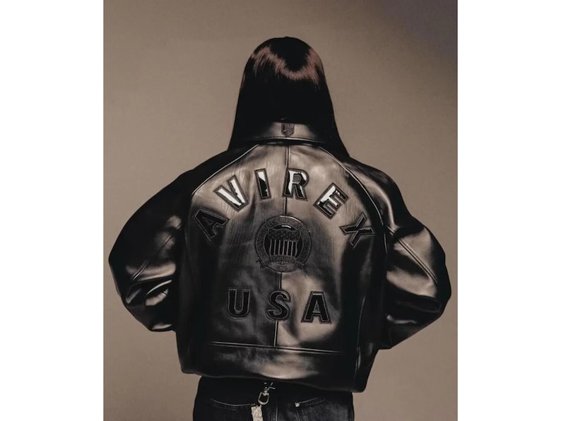 May include: Black leather jacket with the word "AVIREX" and "USA" stitched on the back. The jacket has a round emblem in the center. The jacket is worn by a person with dark hair.