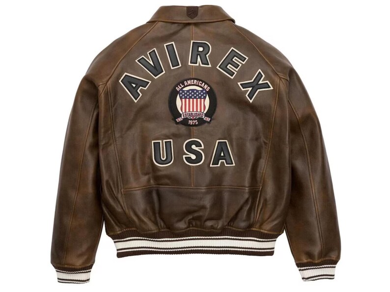May include: Brown leather bomber jacket with the word "AVIREX" arched across the back, above a patch with the American flag and the words "ALL AMERICANS ESTABLISHED 1975". The word "USA" is below the patch. The jacket has white and black striped cuffs and hem.
