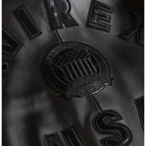 May include: Close-up of a black jacket with raised, glossy lettering and a circular emblem. The emblem features a shield design with the text "AMERICAN" and "ESTABLISHED 1975". The jacket's material appears to be a matte finish.