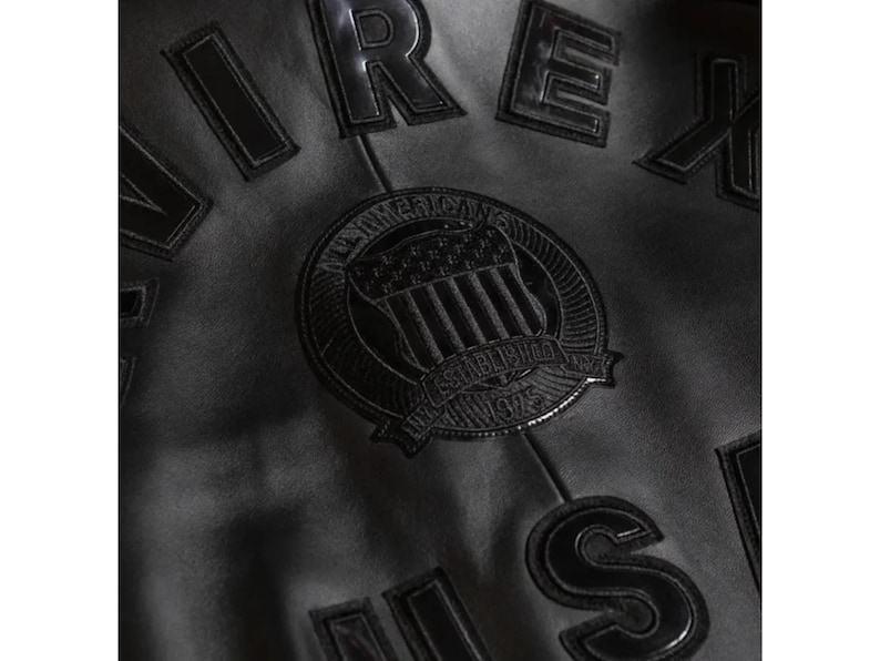 May include: Close-up of a black jacket with raised, glossy lettering and a circular emblem. The emblem features a shield design with the text "AMERICAN" and "ESTABLISHED 1975". The jacket's material appears to be a matte finish.