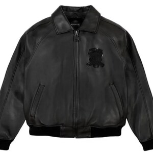 May include: A black leather bomber jacket with a collared neckline and a zipper closure. The jacket features a black emblem on the chest and black ribbed cuffs and hem. The jacket has two front pockets.