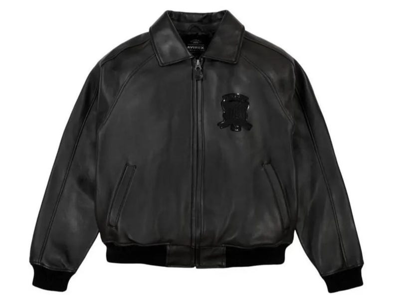 May include: A black leather bomber jacket with a collared neckline and a zipper closure. The jacket features a black emblem on the chest and black ribbed cuffs and hem. The jacket has two front pockets.