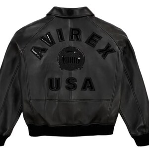 May include: Black leather jacket with the word "AVIREX" arched across the upper back, above "USA". The jacket has a classic bomber style with a ribbed collar, cuffs, and hem. The jacket is a solid black color.