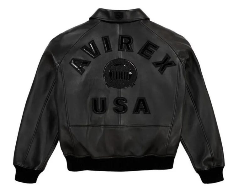 May include: Black leather jacket with the word "AVIREX" arched across the upper back, above "USA". The jacket has a classic bomber style with a ribbed collar, cuffs, and hem. The jacket is a solid black color.