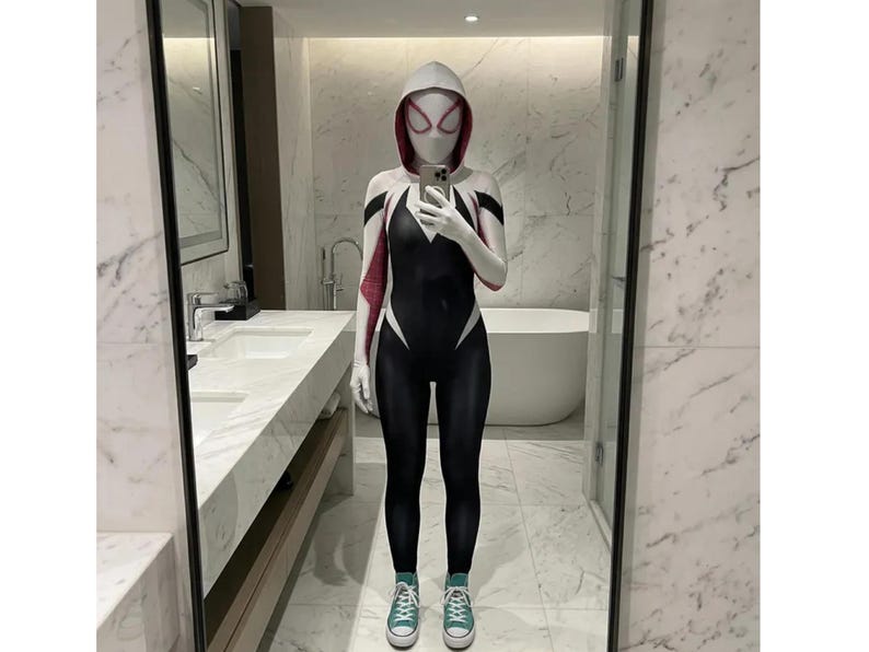 May include: A black, white, and red Spider-Gwen cosplay suit with a hood and mask. The suit has a spiderweb design on the chest and arms. The person is wearing green high-top sneakers. The background is a bathroom with a marble countertop and a bathtub.