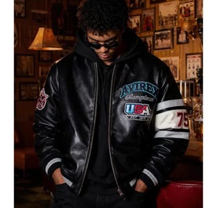 Peut inclure: Veste de bombardier en cuir noir avec le texte "Avirex Champions USA Est. 1975" sur la poitrine. La veste a des poignets ray&eacute;s blancs et noirs et une bande blanche sur la manche avec le num&eacute;ro "75". Un &eacute;cusson &eacute;toile rouge est sur la manche gauche.