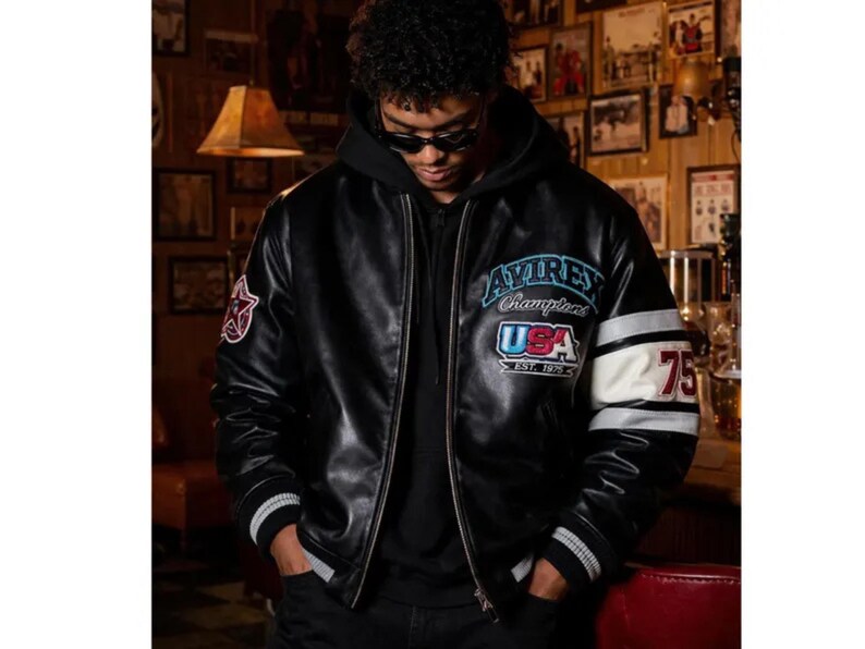 Peut inclure: Veste de bombardier en cuir noir avec le texte "Avirex Champions USA Est. 1975" sur la poitrine. La veste a des poignets ray&eacute;s blancs et noirs et une bande blanche sur la manche avec le num&eacute;ro "75". Un &eacute;cusson &eacute;toile rouge est sur la manche gauche.