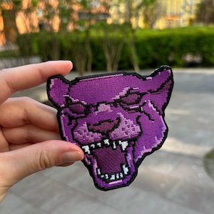 May include: A purple embroidered patch depicting a stylized panther head with an open mouth and visible teeth. The patch has a black border and is held against a blurred outdoor background.